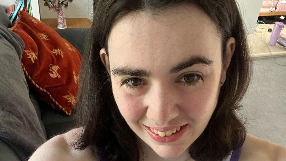 Lucy looking at the camera and smiling in a selfie. There is a grey sofa behind her with a cushion on it.
