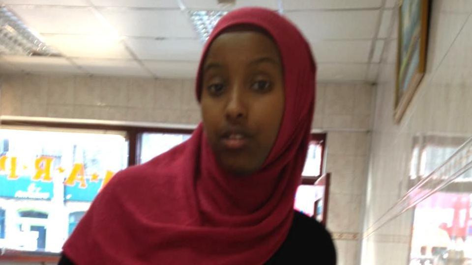 Zahwa Salah Mukhtar in a red headscarf and black top. She stands inside a building with cream tiles on the walls and a window at the back. 