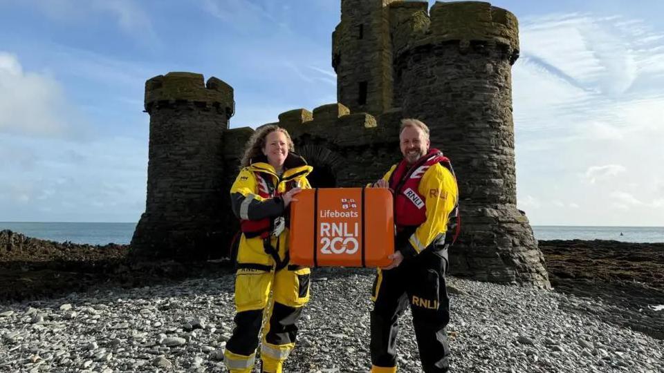 Two RNLI crew, a man and a woman, wearing their protective yellow suits and red lifejackets. They are both holding onto an orange fiberglass case with the writing "RNLI 200" on the side, which contains the scroll. They are standing in front of a small, castle-like building on an island in the sea.