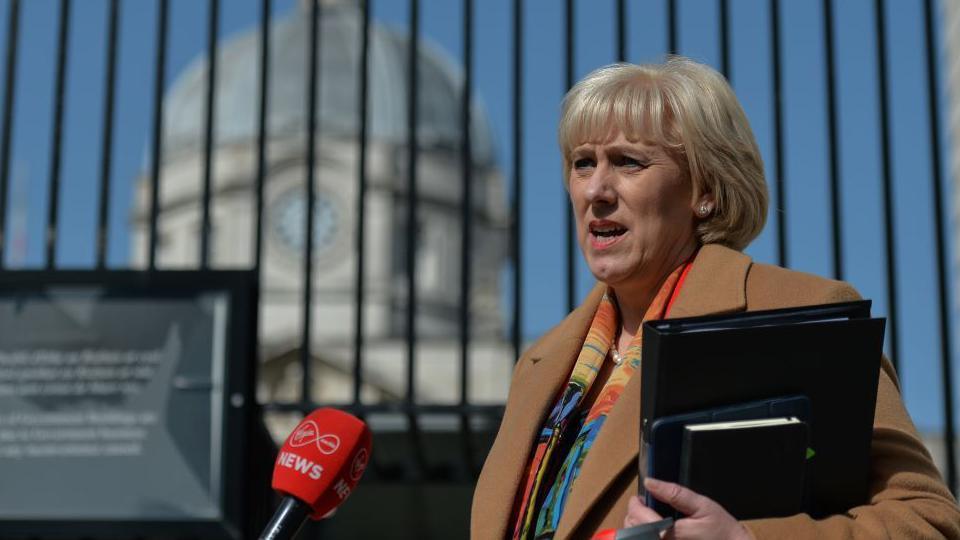 A woman with short blonde hair and fringe wearing a brown coat and multi-coloured shirt talks into a mic as she holds a black file and tablet case.