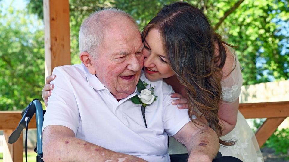A woman in a wedding dress on the right smiles as she looks at a man. The elderly man is wearing a white roses pinned to to his top and is sitting in a wheelchair with the handle visible on the left.