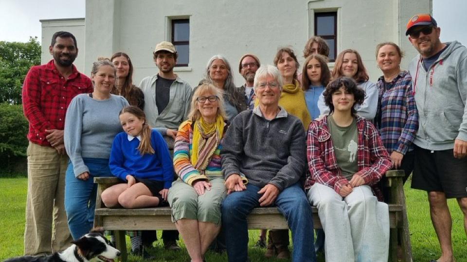 A group of 16 men, women and children stand in front of a large white house. All are smiling and one man looks happily down at a dog. In the centre of John and Nikki who are founder members.