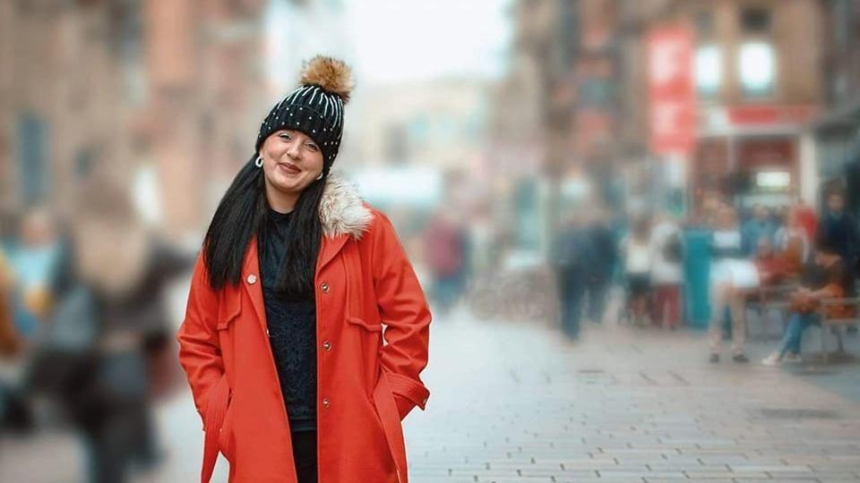 A photo of Lisa looking directly at the camera as she stands outside on a pedestrian street. The background is blurred out and Lisa's full length red coat stands out from the background. She has a black bobble hat on her head and has long dark hair. She is smiling at the camera.