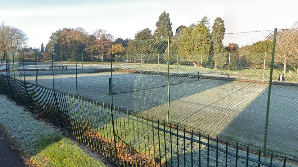 A green astro tennis court surrounded by sports chainmail fencing. There are torso high spiked metal railings in the foreground next to a footpath, with the rest of the park, including mature trees in the background.