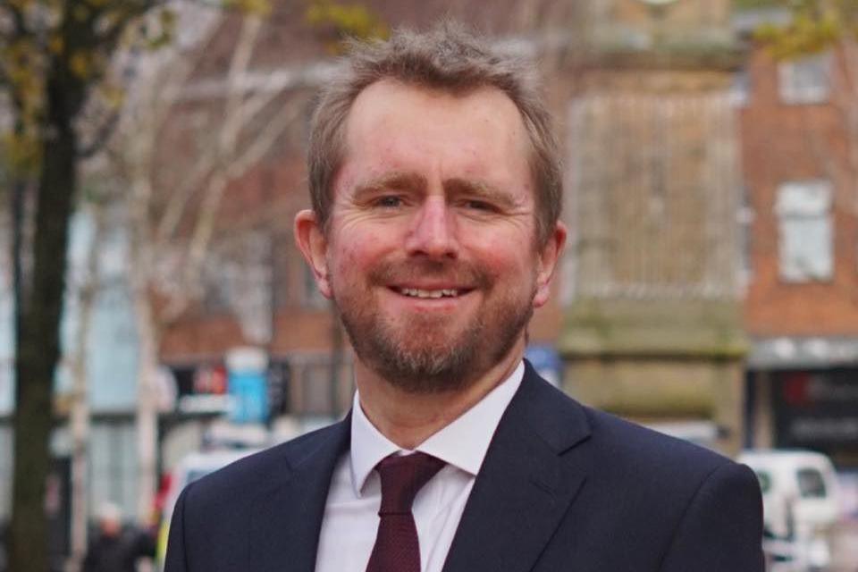 A man stands outside in a street. He has dark blonde hair and a beard. He is wearing a dark blue suit jacket, a white shirt, and a maroon tie. The street in the background has shops on the ground floor.