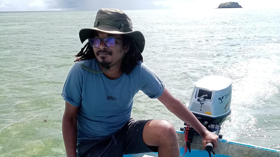 A man with  dark hear, short beard, a floppy hat, glasses and a blue T-shirt steers a motorboat