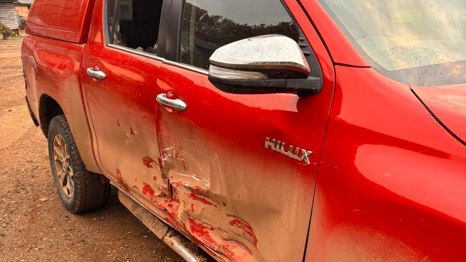 A red Hilux pickup, a famously strong vehicle, with a contorted front and back door, a bent chassis, and a smashed window.