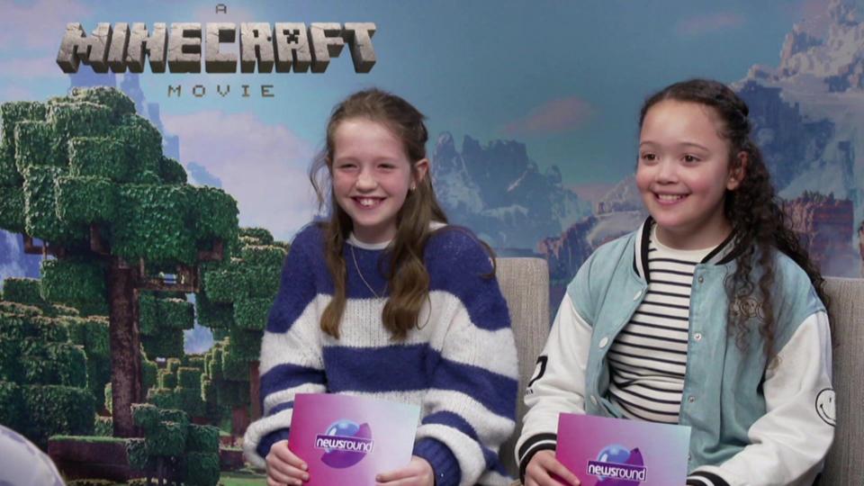 Two girls smiling in front of a Minecraft Movie backdrop, they are holding Newsround cue cards.