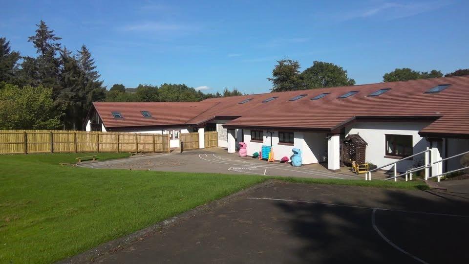 A single storey school building with a long sloping roof and a white facade. There is a large area of lawn in the front and a wooden fence.