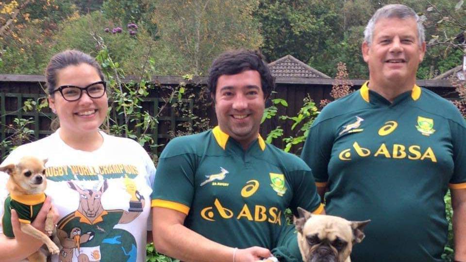 A woman and two men wearing South African rugby shirts, holding a chihuahua and a pug standing outside in a garden.