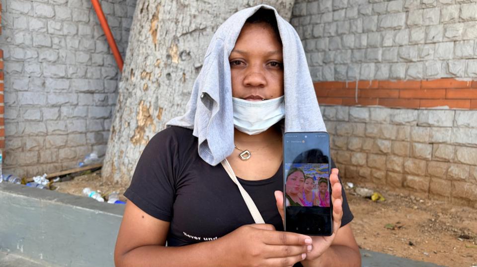 Deilisbeth Herreira stands looking at the camera with a T-shirt on her head and a face mask resting over her chin. She is holding up a photo of her daughters on a phone.