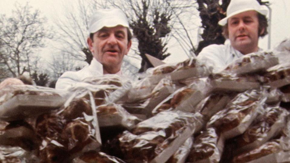 Two men in baker's uniforms stood behind a huge pile of hot cross buns on a market stall.