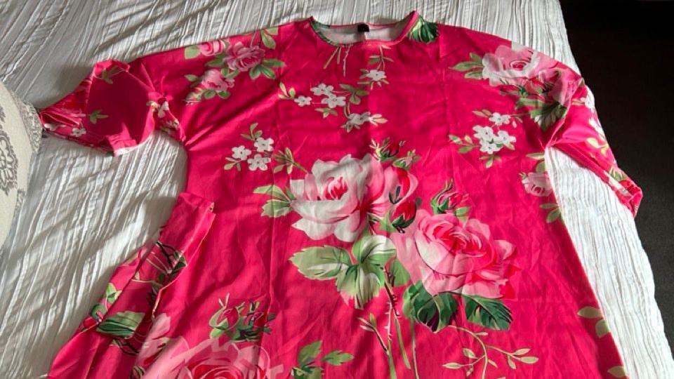 A pink short sleeved dress with floral patterns is laid out on top of a white cloth background. 