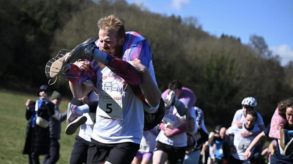 A group of people carrying others on their shoulders. Some are giving the person they are carrying a piggy back, others are carrying with the person's feet in front of them, and them hanging down their back.
