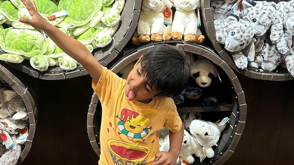 Young boy with his tongue stuck out. He is posing with his right arm in the air and left arm on his side. He is wearing a yellow t-shirt with a cartoon character on it. He is in front of barrels of cuddly toys. 