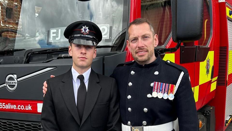 Ben and Steve Wright stood in front of a fire engine. Ben is wearing a suit and hat and Steve is wearing a firefighter's dress uniform, with medals on it.