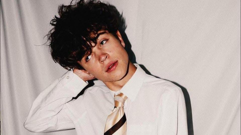 Tom A Smith, in white shirt and a gold and black tie, stands facing the camera with his head tilted and his right arm pressed towards his right ear, with a large mop of dark, tousled hair