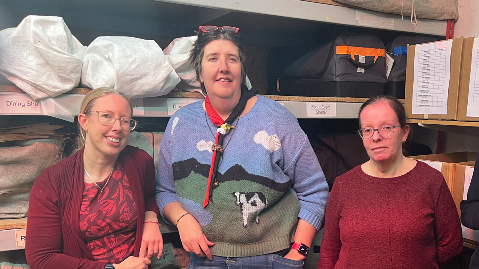 A woman with blonde hair and glasses, a taller woman with dark hair wearing glasses on her head, and a third woman wearing glasses wearing a red jumper standing in front of shelving