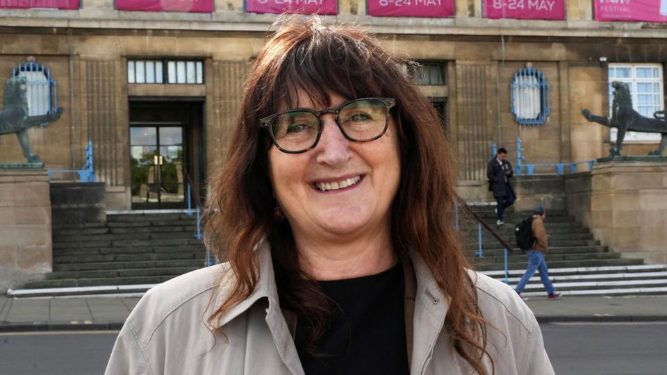 Lucy Galvin, Green leader of Norwich City Council standing outside of City Hall. She is looking directly at the camera and is smiling. She is wearing a black T-shirt with a grey jacket over the top and has brunette hair and a pair of brown framed glasses over her eyes.