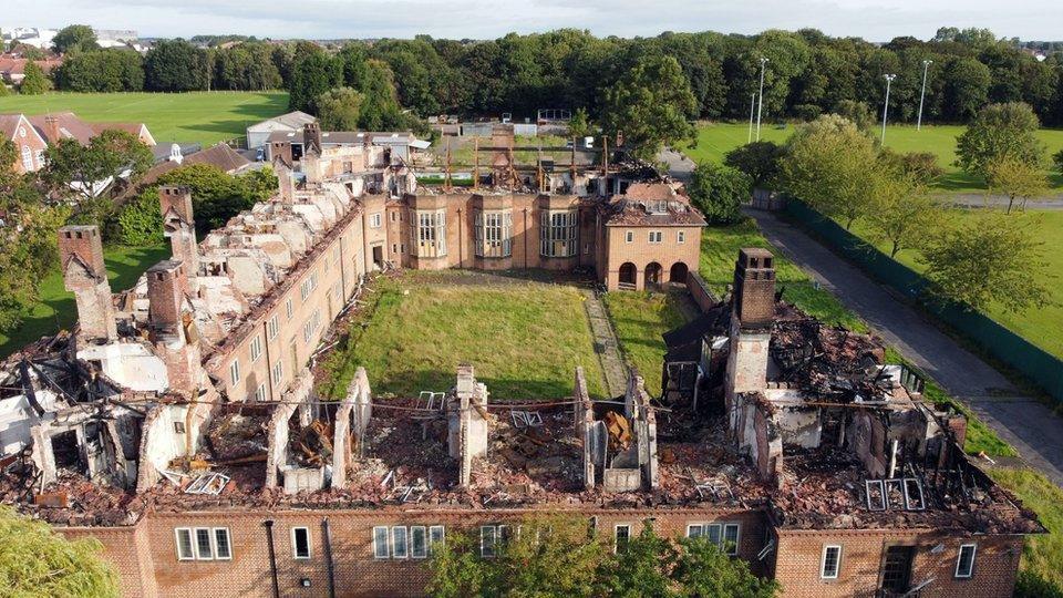 An aerial image of Henderson Hall after the fire. Its roof has been completely destroyed. Rubble has been left on the floor of the exposed upper floors. In the background is parkland and trees.