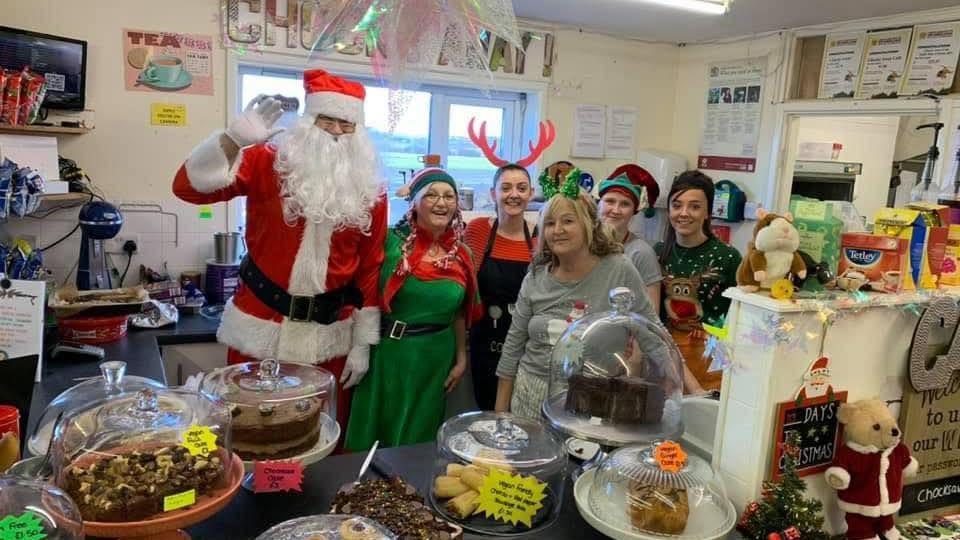 A group of cafe staff in Christmas fancy dress behind a counter filled with cakes. Someone dressed as Santa stands next to them, some of the staff have reindeer antlers on their heads. In front of them are various sweet treats under glass lids.