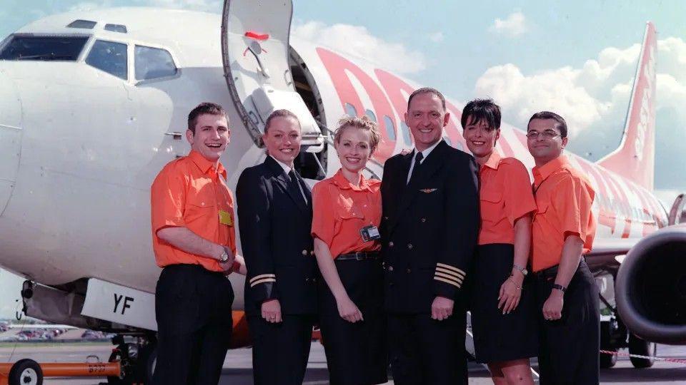 Six crew members including a man and woman wearing pilot's uniforms stand in a group outside an Easyjet plane. Four of them wear bright orange short sleeved shirts and black trousers or skirts. They are all smiling.