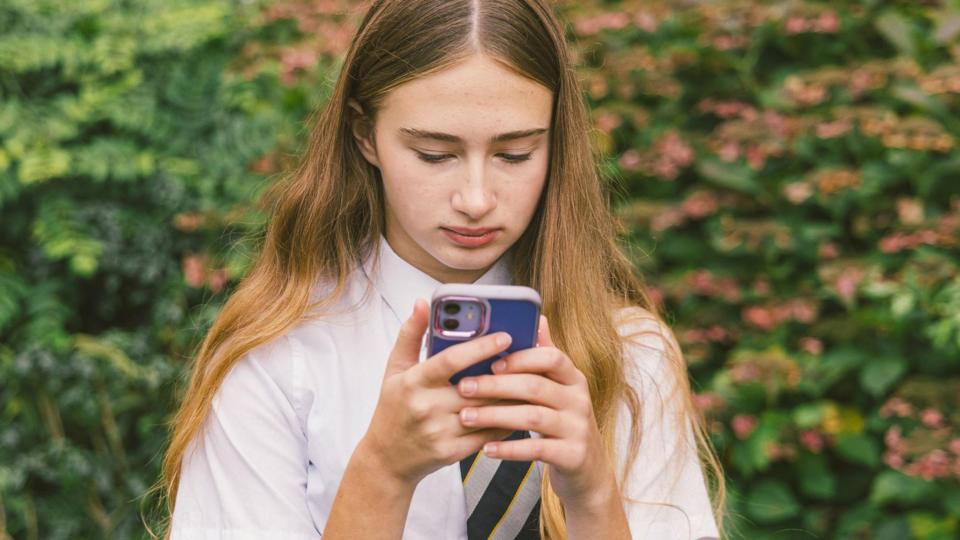 A teenage girl with long blonde hair wearing a school shirt and tie is looking at her mobile phone, which has a blue cover.