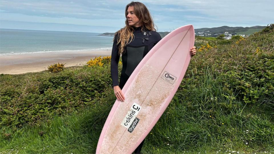 Lucy Campbell wearing a black wetsuit and holding a pink surfboard up. She is standing on grass high above the sea and sand in the background.