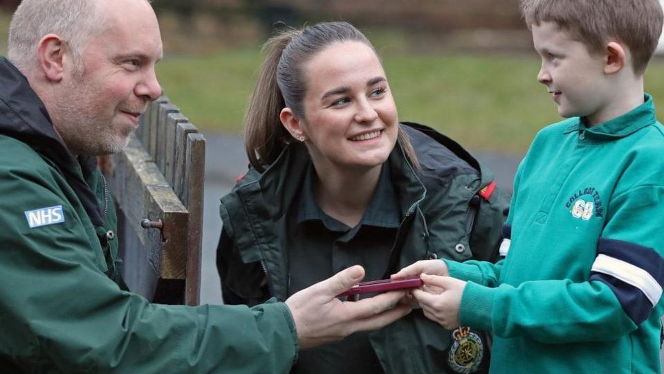 A male emergency service worker and female emergency service worker facing Tyler, who is 8 years old, outside. The workers are pictured in green NHS uniforms while Tyler is pictured wearing a blue sweatshirt.