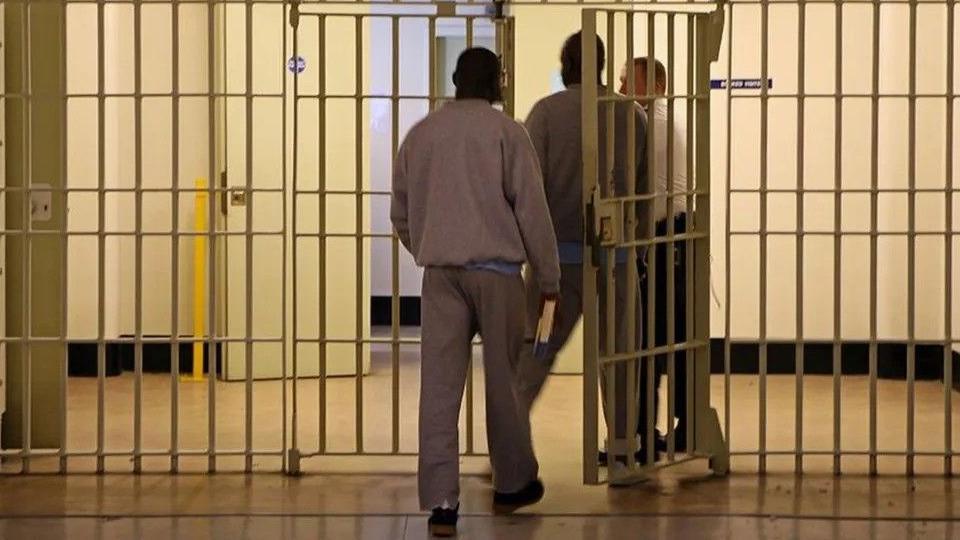 A prison officers lets two prisoners, both wearing grey tracksuits, through a barred door into another section of the prison