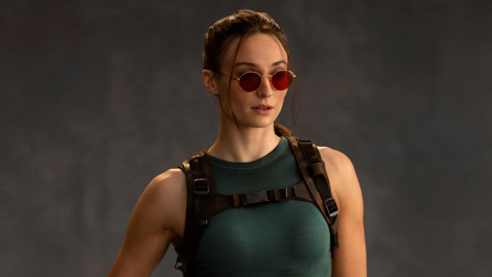 A photo shows Sophie Turner as Lara Croft. She wears red-tinted sunglasses, her hair tied back. She wears a green vest top and tight, thigh-length shorts. One of her fingerless-gloved hands rests on a pistol in a thigh holster.