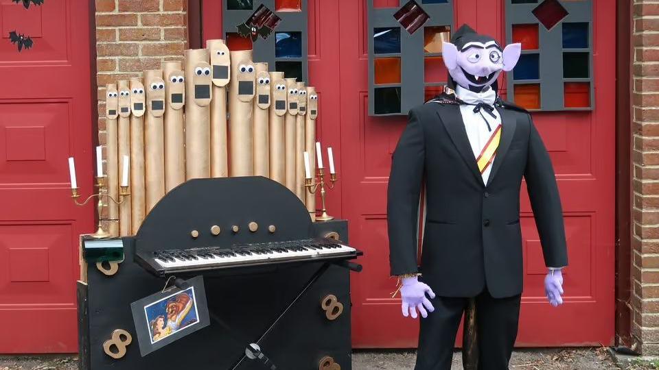 A scarecrow of Count von Count from Sesame Street. He is purple and stood next to an organ.