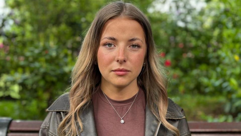 Close-up portrait of Ellie sitting on a park bench, wearing a brown top and dark jacket, with shoulder-length hair and a small pendant necklace, with green foliage blurred in the background.