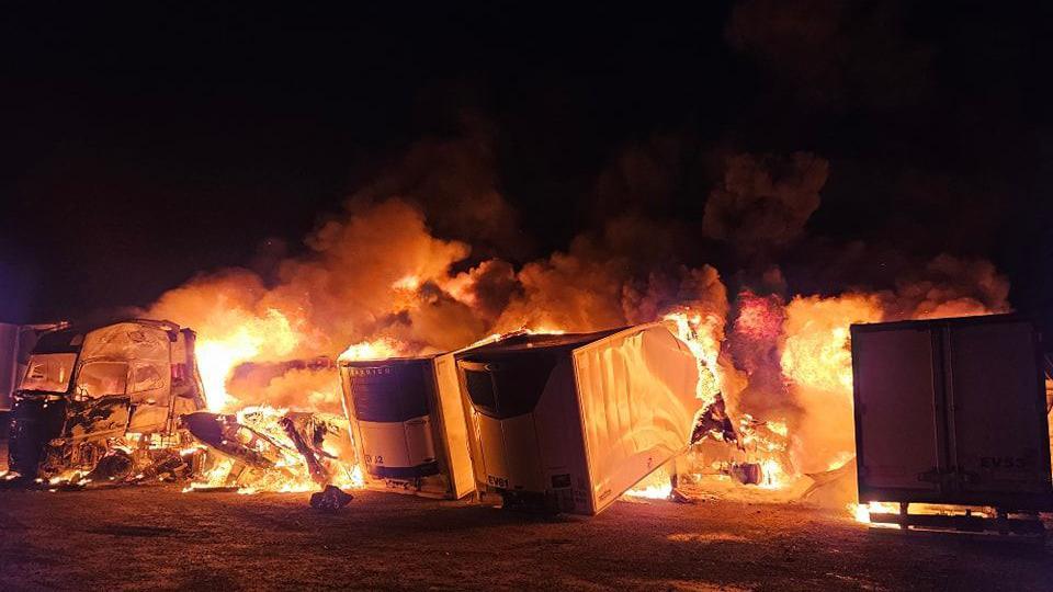 Four lorries are completely consumed in bright orange flames, with black smoke billowing from them