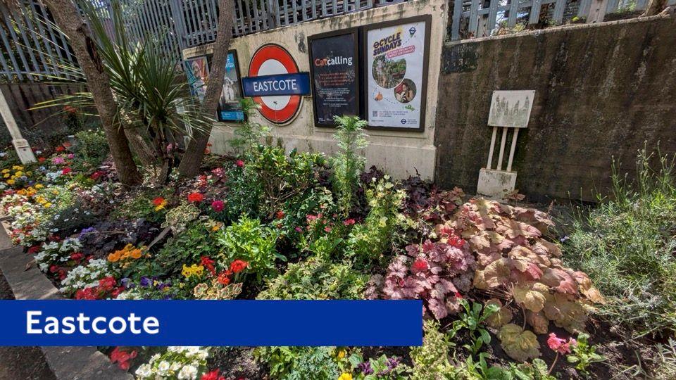 The Eastcote station roundel is in the background and in front is a full flowerbed of colourful plants and flowers.