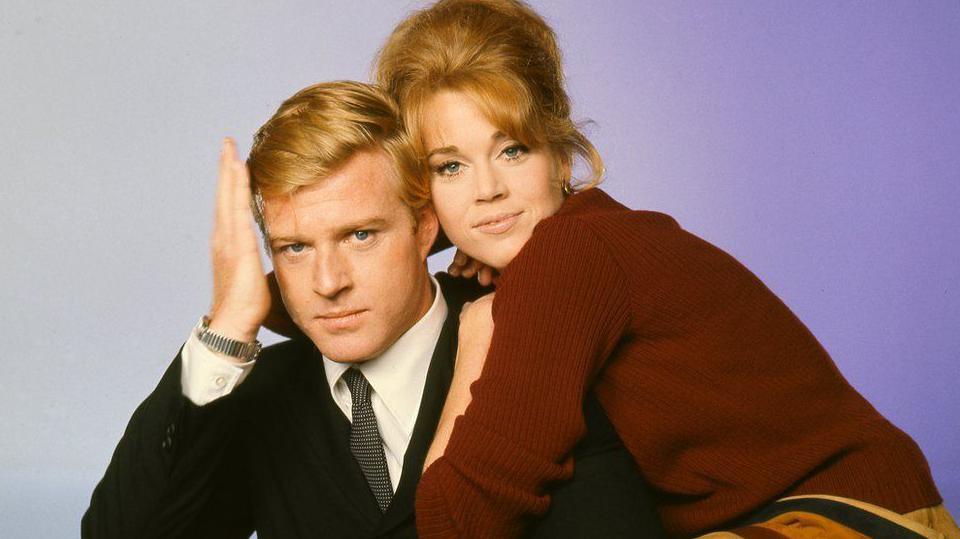 Robert Redford and Jane Fonda in a colour promotional photo for Barefoot in the Park. Redford is dressed in a suit and is smoothing his hair. Fonda is cuddling him and smiling at the camera.