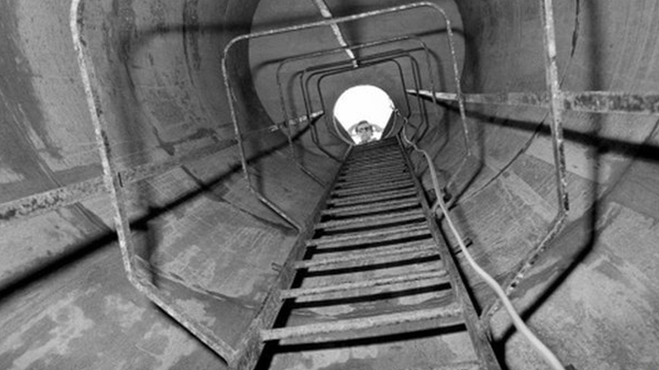 A black and white photograph of a drainage shaft. There is a ladder on the side of the walls and there are railings, leading up to a circle of light at the top through which a person's head can be seen.