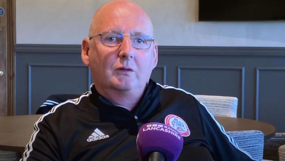 Andy Holt behind a microphone in a function room. He is bald, with glasses and is wearing a black tracksuit top