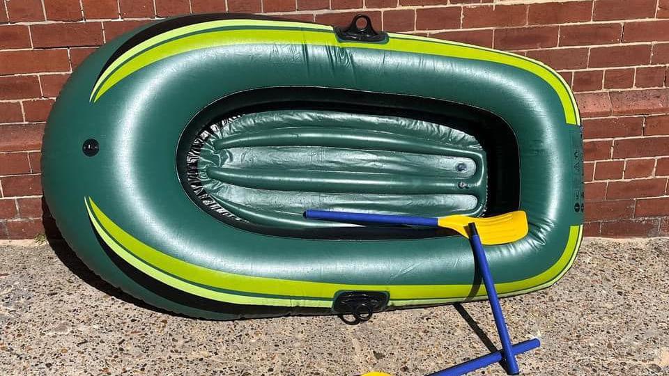 A small inflatable green dinghy with two broken blue oars which has been placed against a brick wall.