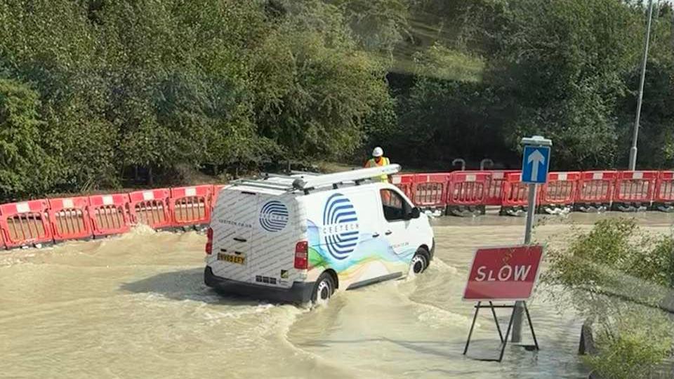 A white van with a ladder on the roof is driving slowly through floodwater. There are red barriers at the side of the road and a red sign with slow written on in white. A workman in a yellow hi-vis jacket and white hardhat is standing near the barriers.