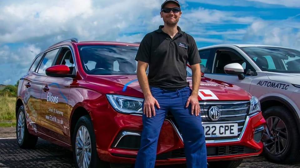 Dan Limond is leaning against the bonnet of a red car. He is wearing a black t-shirt and blue trousers. Dan is smiling and also wearing sunglasses.