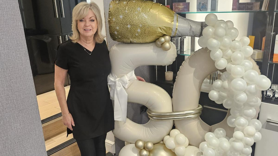 Helen has blonde hair and is wearing a black dress. She is standing next to a display of balloons which features a balloon champagne bottle with small balloon 'bubbles' coming out of it and a big '50' balloon. The balloons are white and gold in colour. There are steps behind Helen which lead to the front door of the salon.