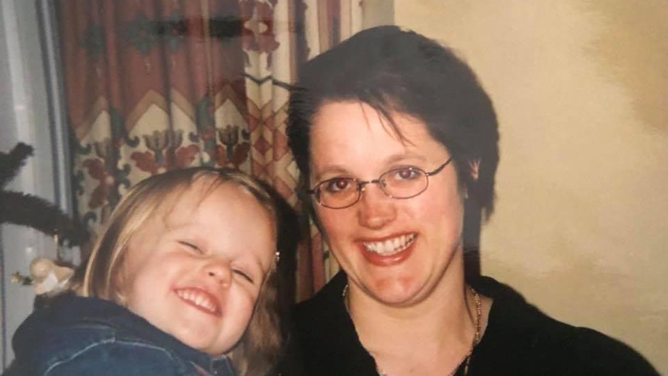 Connie Gibbs is pictured as a child with her mother Zena. Connie is being held by Zena. They are both smiling at the camera. Connie has short blonde hair and is wearing a denim top. Zena has short dark brown hair, she is wearing small glasses, a black top and a necklace.