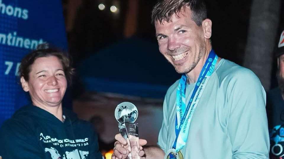 Paul Holborn being presented with his trophy by a woman with dark hair. She's smiling at Paul to the right. Paul is looking off centre and is smiling. He's got dark hair that is wet and is wearing a medal.