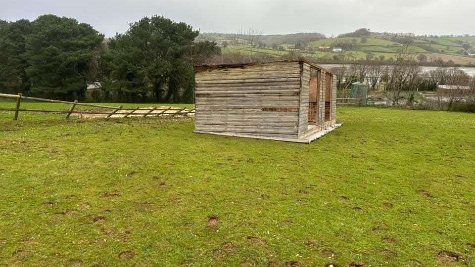 A damaged wooden box shelter on a field. The fence on the left has blown down and there are trees in the background. 