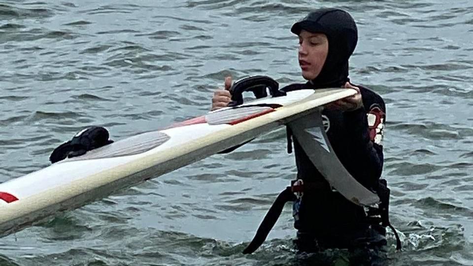 A teenage boy wearing a black wetsuit holds a white windsurfing board as he stands up to the waist in choppy grey water.