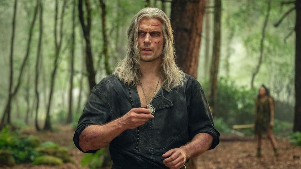 Henry Cavill as Geralt with long grey hair and cuts all over his face. He holds a silver pendant. He's wearing an open-buttoned green shirt with sleeves rolled up. He's in a forest.