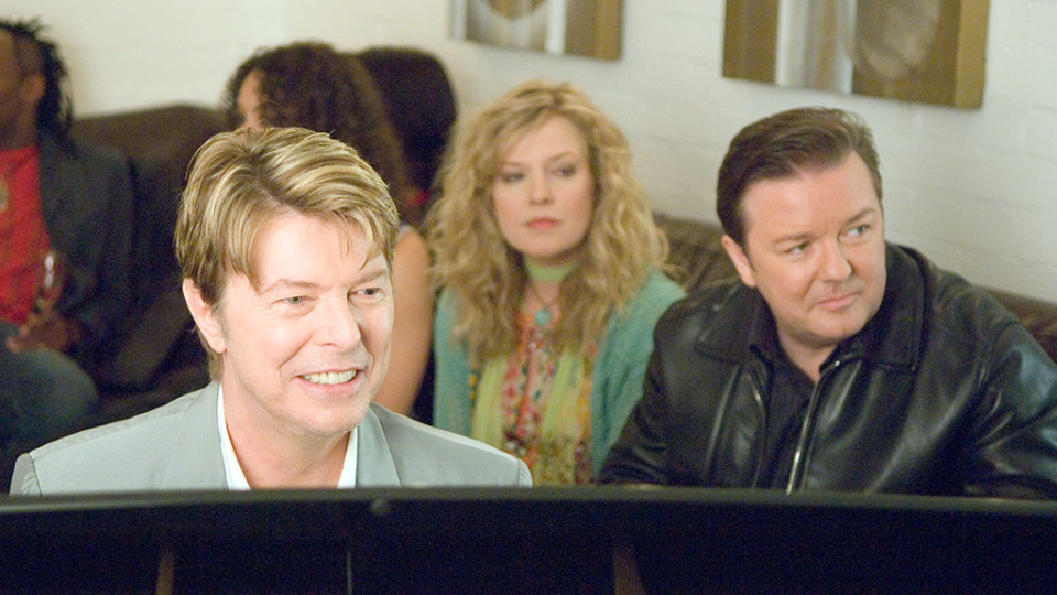 David Bowie and Ricky Gervais sit in a room and look into the distance.