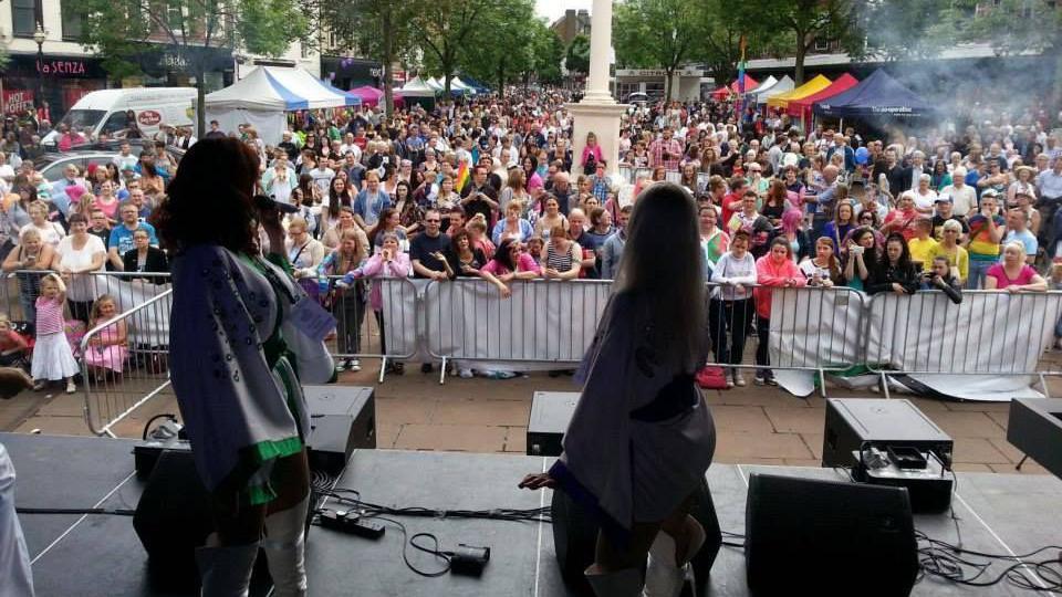 A crowd in front of a stage. People wearing rainbow themed clothes can be seen. Two singers stand on the stage.