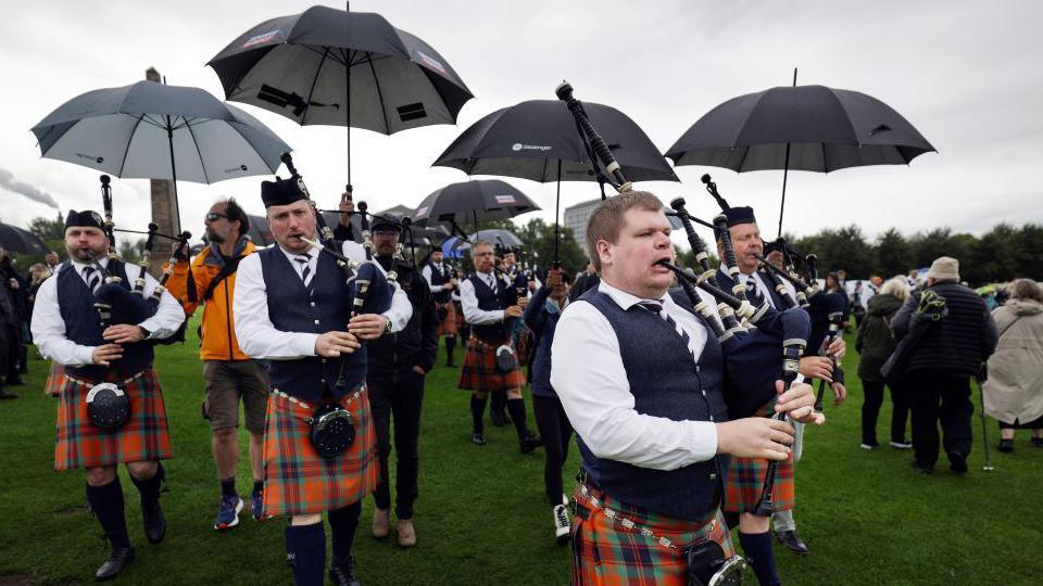Pipers compete while people hold umbrellas over their heads